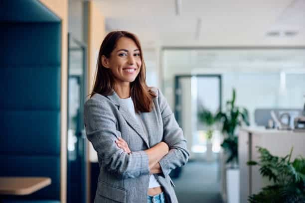 "Confident businesswoman smiling with arms crossed, standing in a modern office environment with glass walls, plants, and natural lighting. She is wearing a grey blazer over a casual outfit."