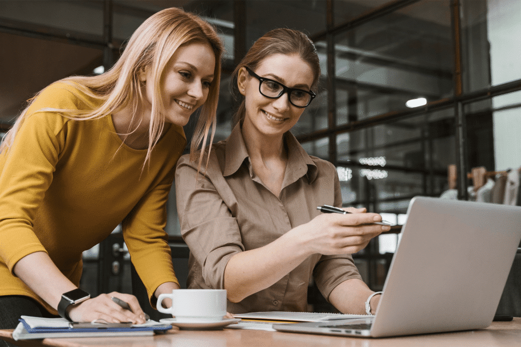 "Two women smiling and working together at a laptop in a modern office setting."